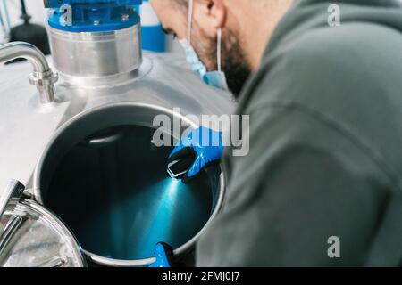 Von oben Seitenansicht eines männlichen Ingenieurs mit blauem Licht Überprüfung von Metallbehältern in der Brauerei während einer Pandemie Stockfoto