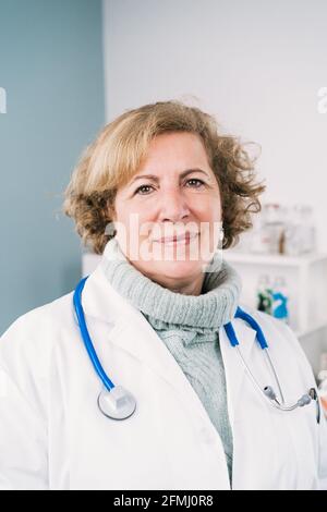 Selbstbewusste Ärztin mittleren Alters in medizinischer Uniform beim Betragen Kamera auf zweifarbigem Hintergrund im Krankenhaus Stockfoto