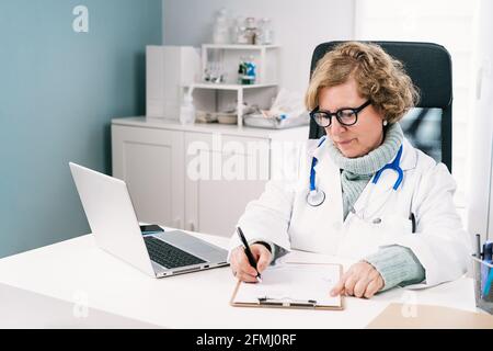 Fokussierte ältere Ärztin in Uniform mit Stethoskop, das Notizen gemacht hat Auf Papier während der Arbeit in der Klinik Stockfoto