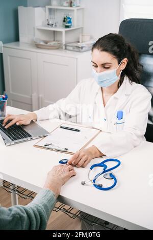 Junge Ärztin mit Laptop, die den Herzschlag der Ernte überprüft Anonymer Patient mit Herzfrequenzsensor am Schreibtisch im Krankenhaus Während einer Pandemie Stockfoto