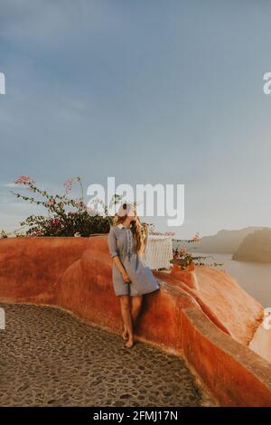 Junge Frau in voller Länge im Sundress, die in der Küstenstadt sitzt Geländer am Ufer und die malerische Aussicht am sonnigen Tag in Santorini Stockfoto