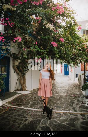 In voller Länge weiblich in stilvollem Kleid berühren blühenden Baum rosa Blumen, während sie auf einer Fußgängerstraße in der Nähe der schwarzen Katze stehen In kleinen authentischen Stockfoto