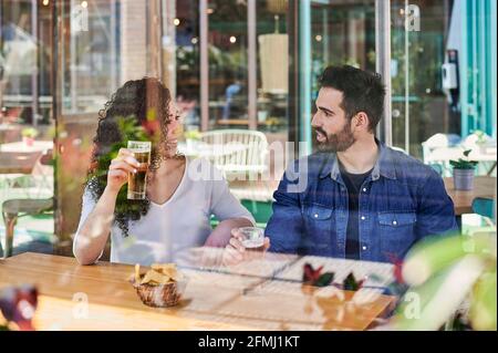 Durch das Glas fröhliches ethnisches Paar mit Gläsern Bier Und Kartoffelchips reden, während sie sich gegenseitig angucken Sonnenlicht Stockfoto