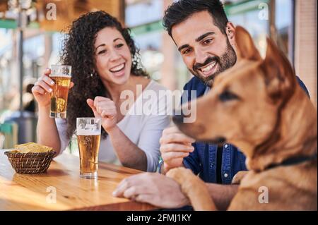 Fröhliches ethnisches Paar mit Gläsern Bier und Kartoffelchips Im Sonnenlicht gegen reinrassigen Hund am Tisch sprechen Stockfoto