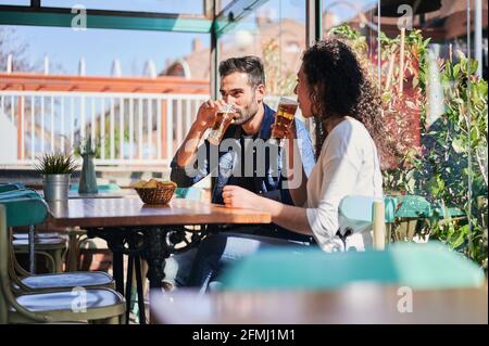 Fröhliches ethnisches Paar, das Bier mit Kartoffelchips trinkt Restaurant in Sonnenlicht Stockfoto