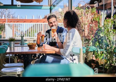 Fröhliches ethnisches Paar mit Gläsern Bier und Kartoffelchips Reden, während man sich im Sonnenlicht ansieht Stockfoto