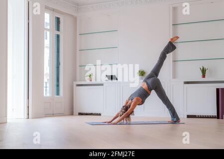 Seitenansicht des friedlichen weiblichen Gleichgewichts in Tri Pada Adho Mukha Svanasana, während man auf der Matte steht und Yoga praktiziert Zimmer zu Hause Stockfoto