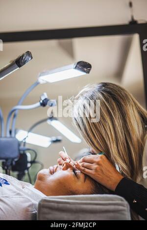 Seitenansicht eines nicht erkennbaren Kosmetologen mit einer Pinzette, die gefälschte Wimpern aufführt Für die Erweiterung auf das Auge des ethnischen Kunden im Salon Stockfoto