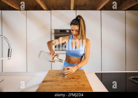 Sportlerin für Erwachsene im aktiven Ohr mit Pferdeschwanz, der Wasser aus der Flasche in das Glas am Tisch im Haus gießt Stockfoto