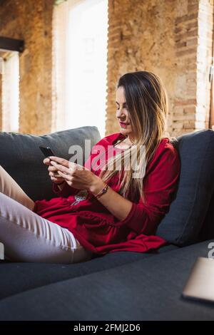 Weibliche Remote-Mitarbeiter SMS-Nachrichten auf dem Mobiltelefon, während sie sitzen Couch gegen Tablet im Loft-Stil Haus Stockfoto