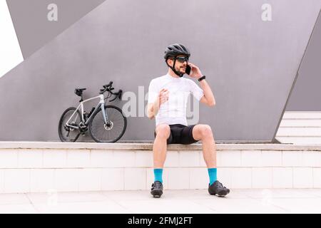 Bärtiger männlicher Biker in Sportkleidung und Sonnenbrille auf dem Bürgersteig sitzend Beim Gespräch mit dem Handy und dem Blick auf das Fahrrad Stockfoto