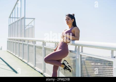 Seitenansicht der jungen sportlichen Sportlerin mit erhobenem Bein Blick weg auf Brücke in Sonnenlicht Stockfoto