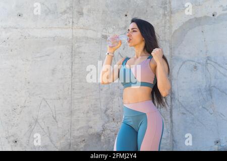 Durstige Sportlerin in Sportkleidung Trinkwasser aus der Flasche Nach dem Arbeiten, während Sie auf grauen Hintergrund wegschauen Stockfoto