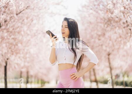 Junge, zärtliche Frau mit der Hand auf der Hüfte, die eine Sprachnachricht sendet Auf dem Mobiltelefon, während Sie bei Tageslicht wegschauen Stockfoto