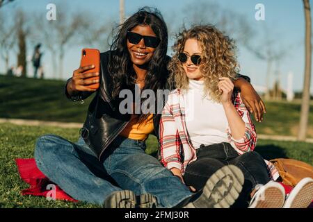 Fröhliche multirassische weibliche beste Freunde umarmen im Frühlingsgarten und Selbstschuss auf dem Smartphone am sonnigen Tag Stockfoto