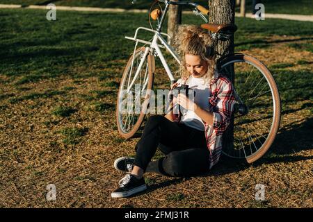 Inhalt Frau sitzt in der Nähe des Fahrrads im Garten und schaut durch Bilder auf professionelle Fotokamera Stockfoto