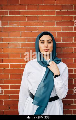 Muslimische Frau mit traditionellem Kopftuch, die in der Stadt gegen eine Mauer steht Und Blick auf die Kamera Stockfoto