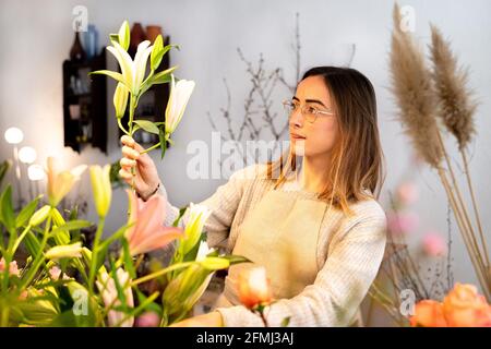 Konzentrierte junge weibliche Floristin in Schürze und Brillen Anordnung duftend Blumen in Vase bei der Arbeit im Blumengeschäft Stockfoto