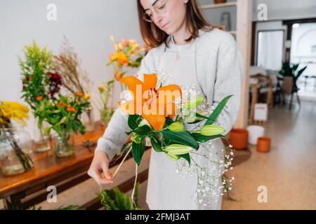 Fokussierte junge ethnische weibliche Floristin in lässiger Kleidung und Schürze Zartes Bouquet von oranger Lilie und weißer Gypsophila einbinden Blumengeschäft Stockfoto