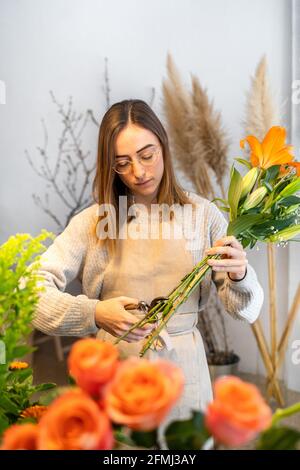 Fokussierte junge ethnische weibliche Floristin in lässiger Kleidung und Schürze Schneiden von Stengeln mit frischen, hellen Blüten und Schnittern Blumengeschäft Stockfoto