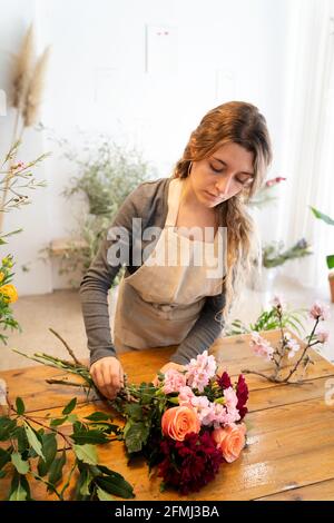 Konzentrierte junge weibliche Floristin mit langen welligen Haaren in casual Kleidung und Schürze arrangieren eleganten Strauß von Rosen und sortiert Blumen im Laden Stockfoto