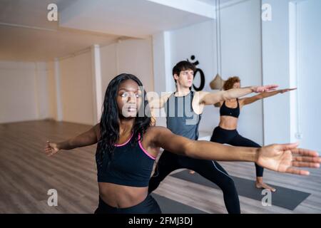 Afroamerikanische Frau in Gesellschaft verschiedener Menschen, die Yoga in Warrior Two Pose im Studio praktizieren Stockfoto