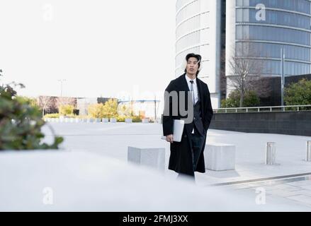Junge ethnische männliche Führungskraft im Mantel mit Hand in der Tasche Und Netbook, das beim Bummeln in der Stadt wegschaut Stockfoto