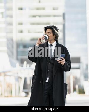 Junge gut gekleidete ethnische männliche Führungskraft mit Handy und heiß Trinken Sie, um in die Stadt zu gehen, und schauen Sie weg Stockfoto