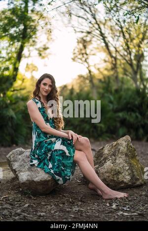 Voller Körper Inhalt junge Frau trägt trendige Maxi Sundress und Lächelnd auf die Kamera blicken, während sie auf einem großen Stein sitzt In einem grünen, sonnigen Wald Stockfoto