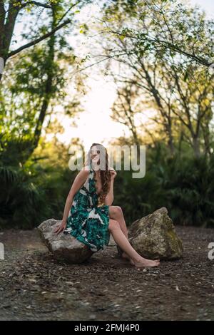 Voller Körperinhalt junge Frau trägt trendige Maxi Sundress berühren Haare sanft und mit einem Lächeln wegschauen, während sie darauf sitzen Großer Stein in grüner Sonne Stockfoto