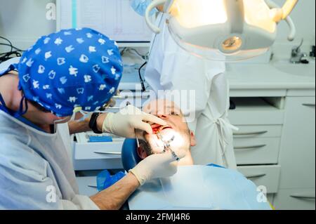 Seitenansicht des anonymen Zahnchirurgen in Uniform und medizinisch Fernglas mit Inhalt Mann auf Stuhl in der Klinik Stockfoto