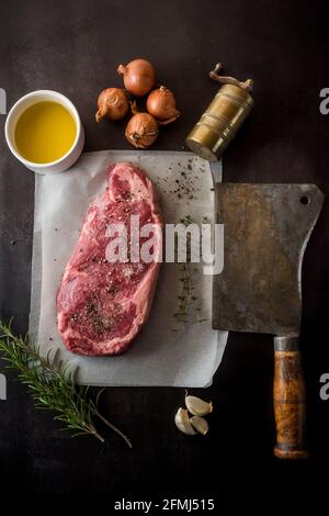 Draufsicht auf ungekochte Fleischstücke mit Gewürzen zwischen frischen Rosmarinzweige und Knoblauchzehen auf dunklem Hintergrund Stockfoto