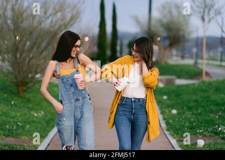 Inhalt beste Freundinnen in lässiger Kleidung zu heißen Getränken In Tumblern berühren Ellenbogen, während sie einander in Stadt Stockfoto
