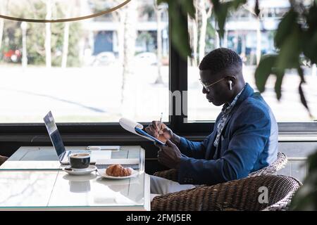 Seitenansicht des konzentrierten afroamerikanischen männlichen Unternehmers beim Schreiben Notizblock, während man mit einem Laptop am Tisch sitzt und darin arbeitet Café Stockfoto