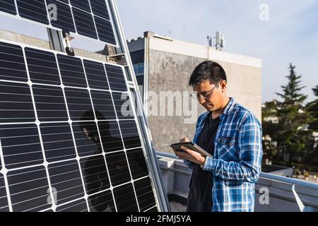 Konzentrierter ethnischer männlicher Techniker im karierten Hemd, der während des Tablet-Besuchs surft Steht in der Nähe von Photovoltaik-Panel in modernen Solarkraftpark gelegen Stockfoto