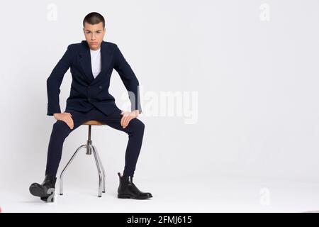 Ernst männliche Modell in modischen Anzug sitzt auf weißem Hintergrund Im Studio und beim Blick auf die Kamera Stockfoto