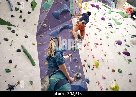 Flache Rückansicht der weiblichen Trainerin, die den männlichen Alpinisten unterstützt Im Klettergurt künstliche Wand im Boulderzentrum Stockfoto