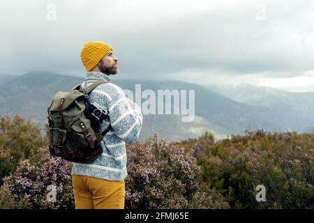 Rückansicht eines nachdenklichen männlichen Campers mit warmer Mütze im Stehen Mit Rucksack im Hochland bewundern und genießen Sie malerische Ausblicke Stockfoto