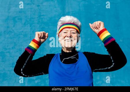 Eine begeisterte ältere Frau in der Sportbekleidung feiert den Sieg mit geballten Fäusten Und erhöhte Arme auf blauem Hintergrund im Studio Stockfoto