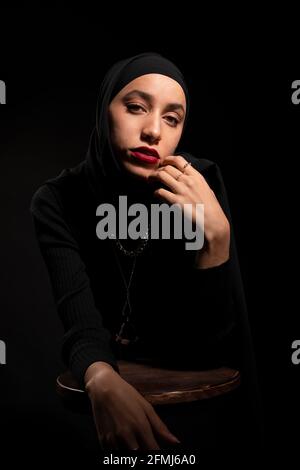 Attraktive junge islamische Frau in schwarzem Outfit und Hijab berühren Gesicht sanft auf Stuhl im schwarzen Studio lehnend und schauend an Kamera Stockfoto