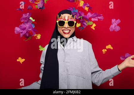 Fröhliche junge muslimische Frau trägt Hijab und lustige Partybrille Stehen vor rotem Hintergrund in der Nähe von fallenden Blütenblättern Stockfoto