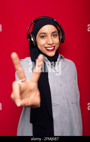 Lächelnd attraktive muslimische Frau in legerer Kleidung und Hijab zuhören Um Lieblingsmusik über Kopfhörer zu hören und mit zwei Fingern dagegen zu zeigen Roter Hintergrund Stockfoto
