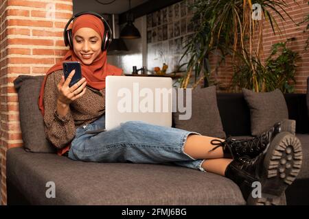 In voller Länge glücklich junge muslimische Frau in bescheidenen Kleidung Hijab Und kabellose Kopfhörer, die auf einer gemütlichen Couch mit Netbook sitzen Runden und smartpho surfen Stockfoto