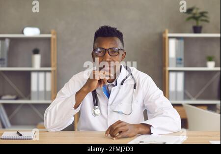 Porträt eines jungen schwarzen Arztes in einer Brille, die am Schreibtisch sitzt Und dem Patienten zuhören Stockfoto