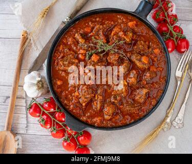 Italienischer Rindereintopf mit Tomaten, Knoblauch, Karotten, Zwiebeln, Petersilie und Sellerie, serviert in rustikaler Eisenpfanne auf Holztischuntergrund Stockfoto