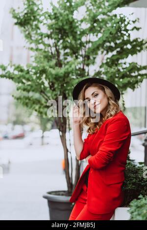 Junge schöne blonde Frau in einem roten Anzug und schwarzem Hut steht im Freien im Hof neben dem Gebäude. Stilvolle Menschen. Weicher, selektiver Fokus. Stockfoto