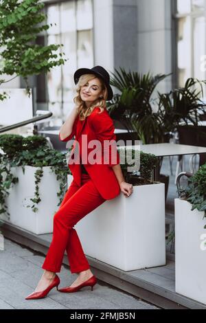 Junge schöne blonde Frau in einem roten Anzug und schwarzem Hut steht im Freien im Hof neben dem Gebäude. Stilvolle Menschen. Weicher, selektiver Fokus. Stockfoto