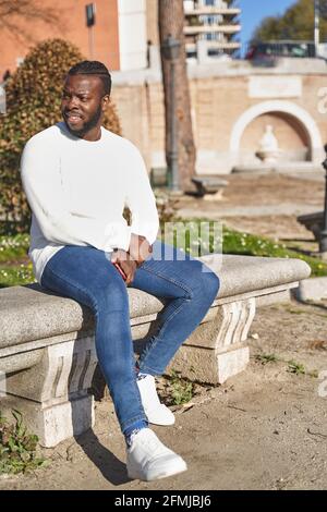 Glücklicher afroamerikanischer Mann, der in einem Park sitzt und Jeans, weißen Pullover und weißen Turnschuhen trägt. Lifestyle-Konzept. Hochwertige Fotos Stockfoto