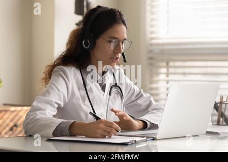 Fokussierte junge Ärztin im Headset, die sich das Webinar ansieht und sich Notizen ansieht Stockfoto
