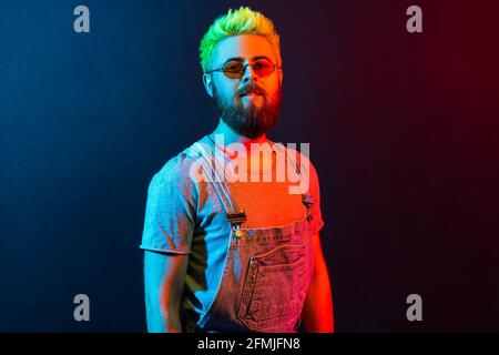 Porträt eines stolzen, hübschen jungen bärtigen kaukasischen Mannes mit grünen Haaren in Denim-Overalls, der mit stolzem und egoistischem Gesicht auf die Kamera blickt. Farbenfroh Stockfoto